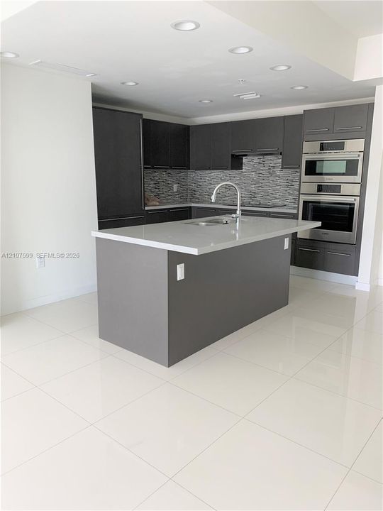 Kitchen featuring dark cabinetry, stone-finish countertops, a central island with an integrated sink, built-in stainless steel double ovens, and polished tile flooring
