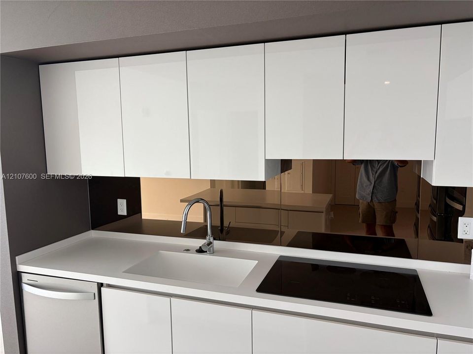 Kitchen featuring high-gloss white cabinetry, white stone-composite countertop, integrated white sink with chrome arched faucet, and a mirrored backsplash