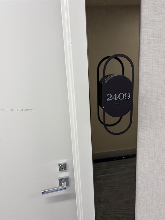 Entry door featuring a metallic lever handle, deadbolt lock, and decorative matte black oval unit identification plaque