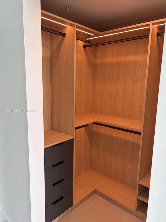 Custom walk-in closet featuring light wood-finish cabinetry, integrated linear LED lighting, dark matte drawers, and multiple hanging rods