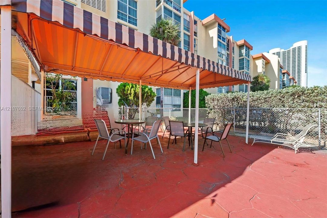Ground-level patio featuring red-toned concrete flooring, a retractable striped fabric awning, and perimeter chain-link fencing