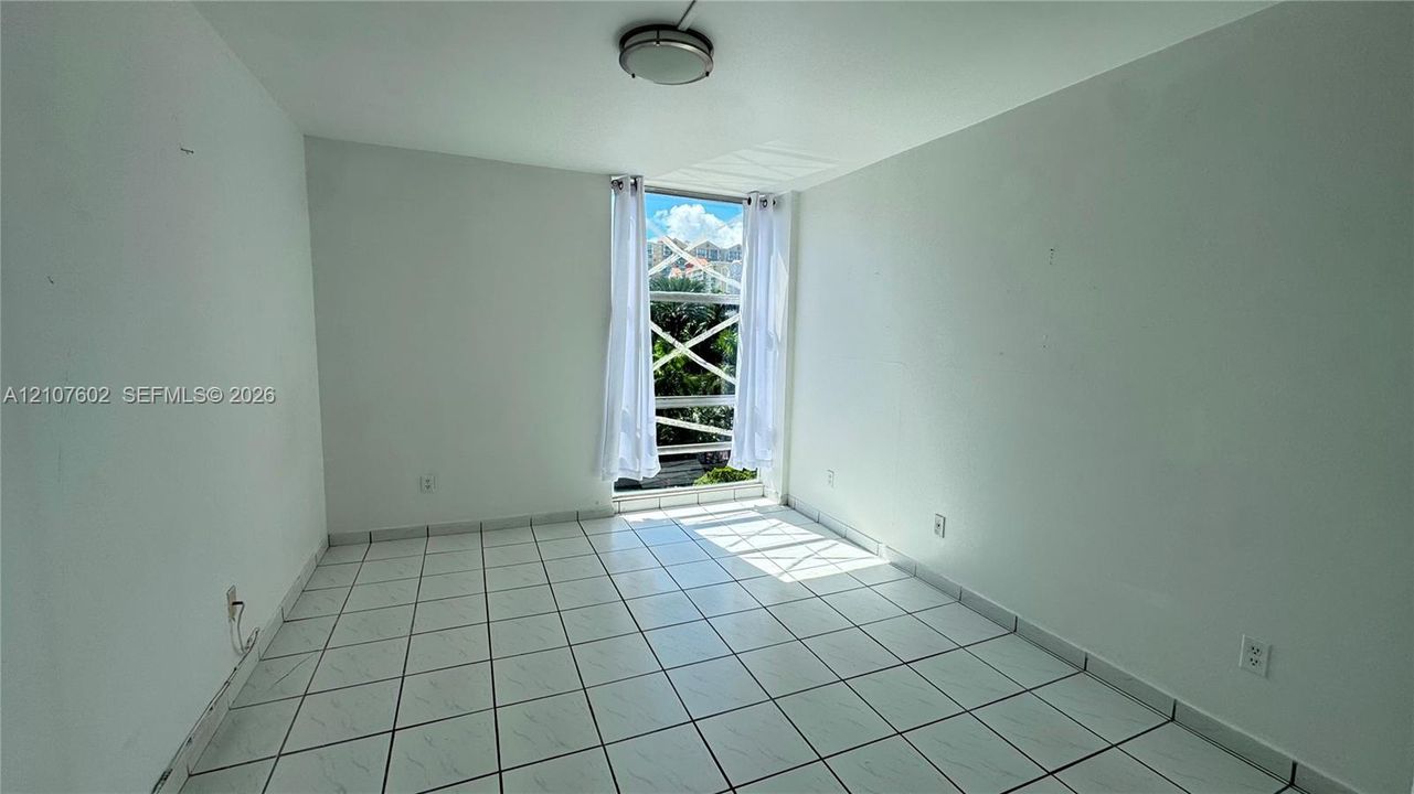Neutral interior room featuring white tile flooring, flush-mount lighting, and a floor-to-ceiling window with a cross-pattern security grille