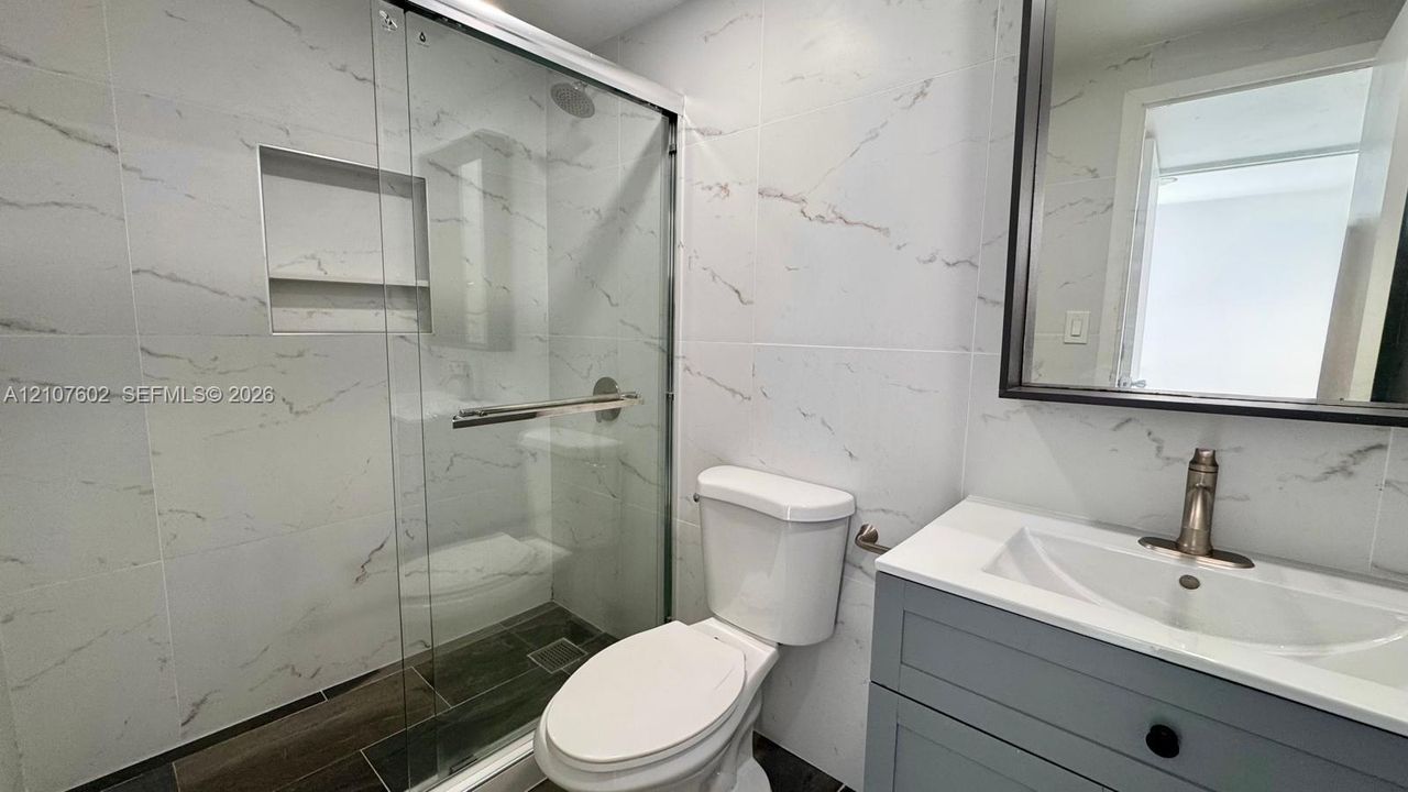 Full bathroom featuring wall-to-wall large-format porcelain tile, glass-enclosed shower with a recessed wall niche, gray shaker-style vanity with an integrated sink, and a framed mirror