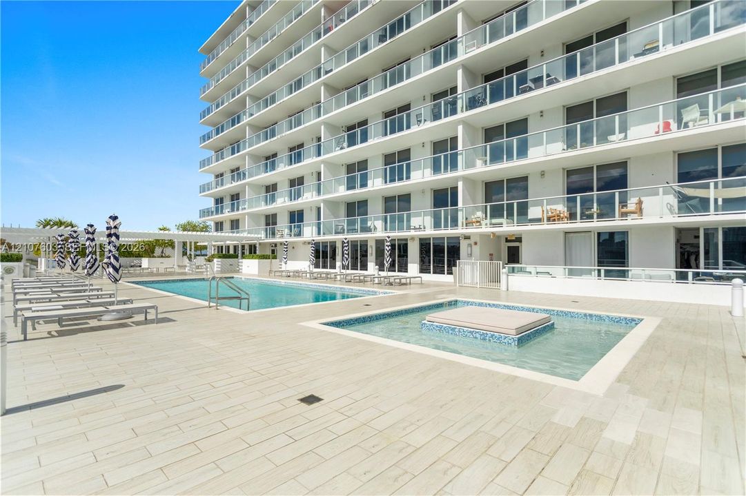 Poolside terrace featuring an in-ground swimming pool, tiled spa, wood-finish porcelain decking, and multi-story building exterior with glass-railed balconies