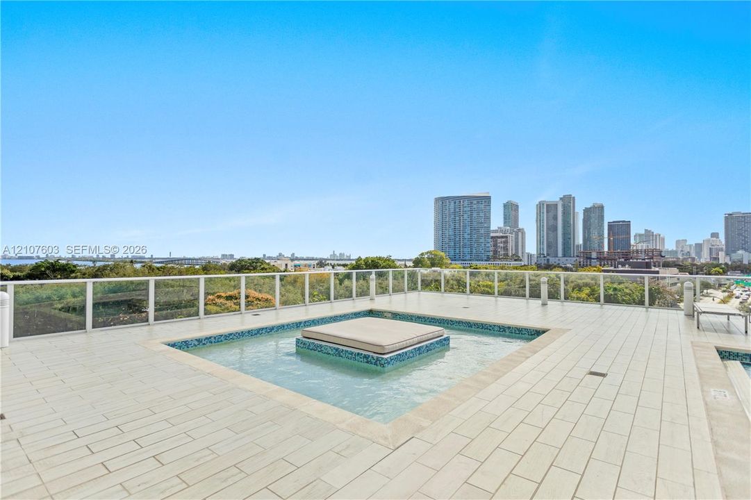 Rooftop terrace featuring large-format tile flooring, a central built-in water feature with mosaic tile accents, a glass panel railing system, and an expansive cityscape backdrop