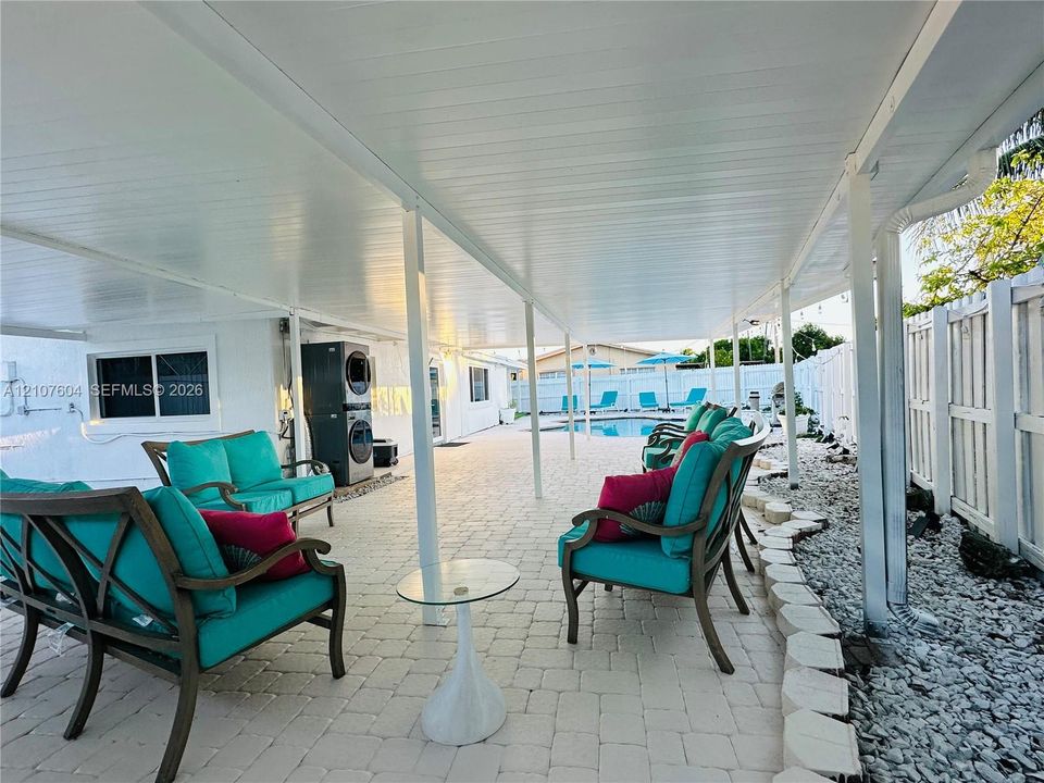 Covered patio featuring paver flooring, a solid roof structure with support columns, white vinyl fencing, and an integrated swimming pool