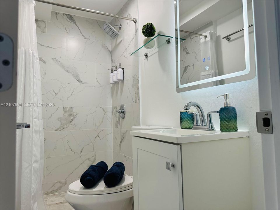Bathroom featuring a shower stall with stone-finish tile surround, a chrome rain-style shower head, a white vanity with an integrated sink, a chrome widespread faucet, and a wall-mounted LED-backlit mirror