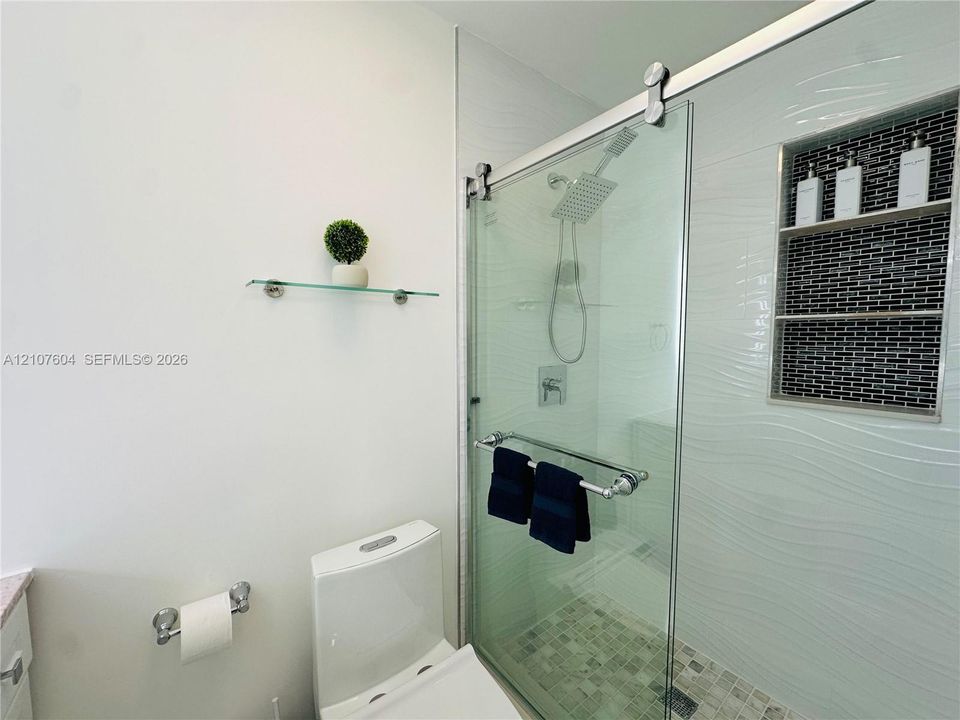 Walk-in shower featuring a sliding glass door, rainfall showerhead, textured tile surround, mosaic stone-finish floor, and a built-in recessed tiled niche