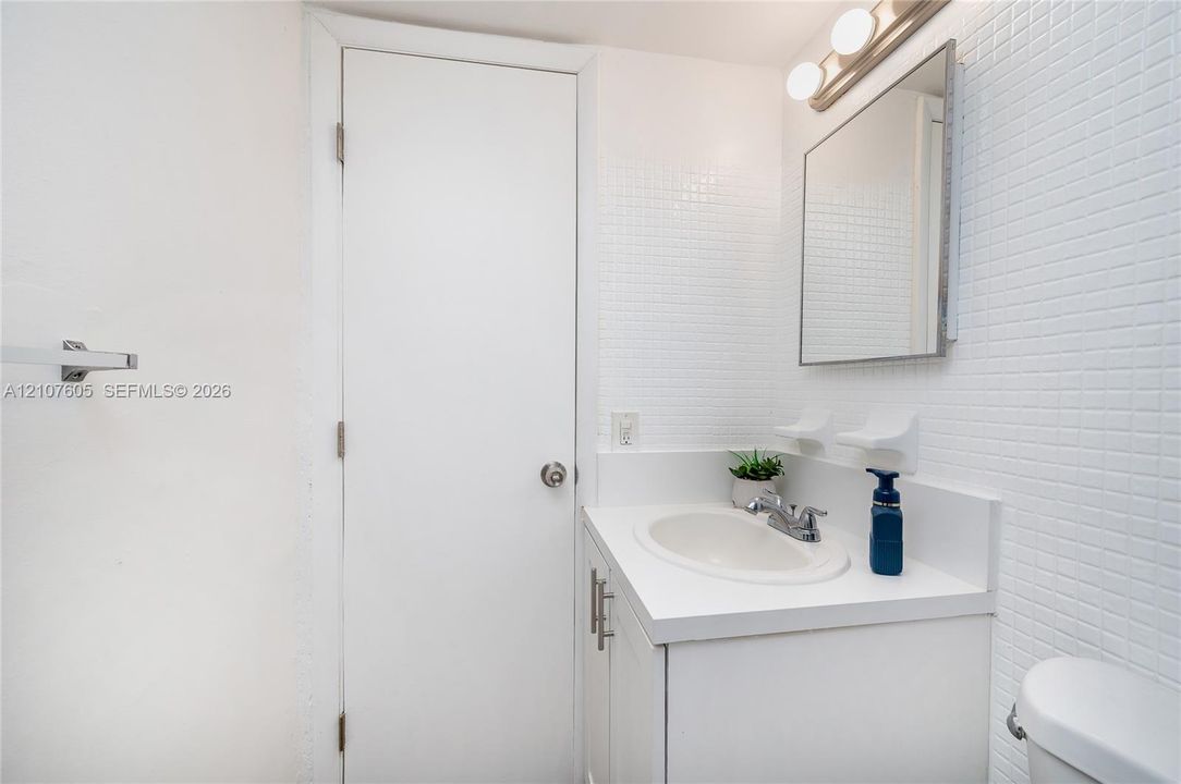 Vanity featuring a white countertop with an integrated basin, square wall-mounted mirror, three-bulb vanity light fixture, and white grid-pattern wall tile