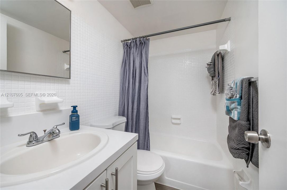 Bathroom featuring a white vanity with a composite countertop and integrated sink, a frameless wall-mounted mirror, white square-tiled wall surround, and a white tub-shower combination