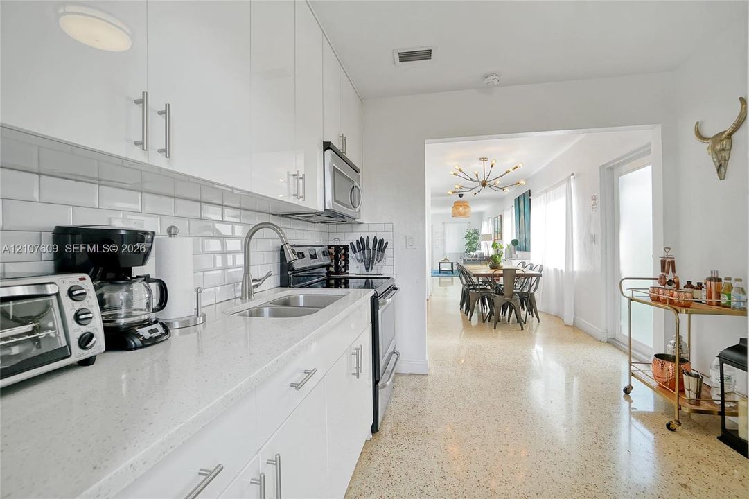 Kitchen featuring white flat-panel cabinetry, light-toned stone-surface countertops, a double-basin stainless steel sink with gooseneck faucet, white subway tile backsplash, and polished terrazzo flooring