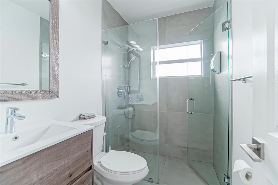 Bathroom featuring a wood-finish vanity with a rectangular basin, glass-enclosed shower with large-format tile surround, integrated shower head system, and chrome hardware