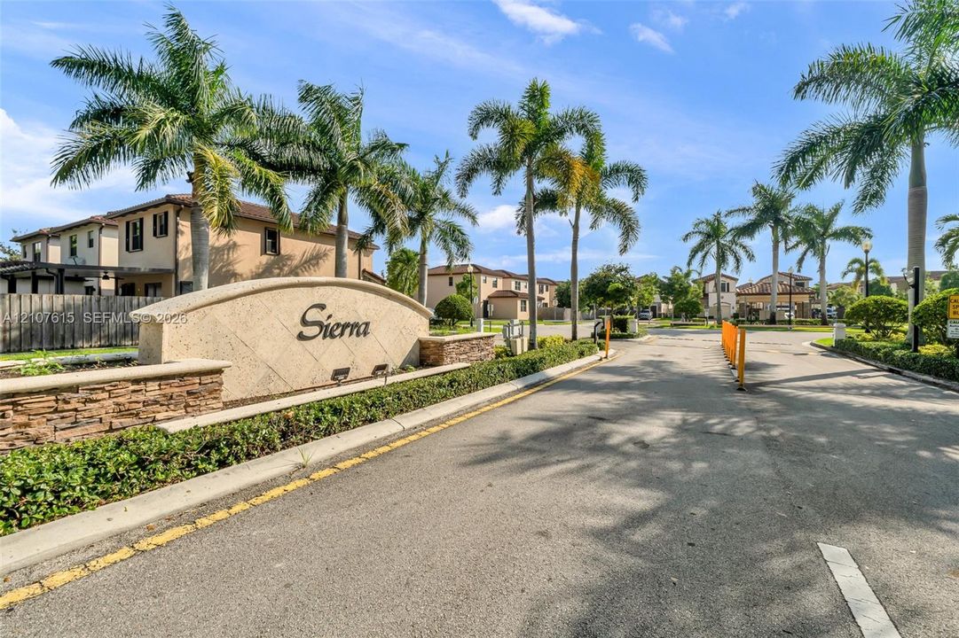 Community entrance featuring a stone masonry monument sign, asphalt paved roadway, manicured landscaping, and mature palm trees
