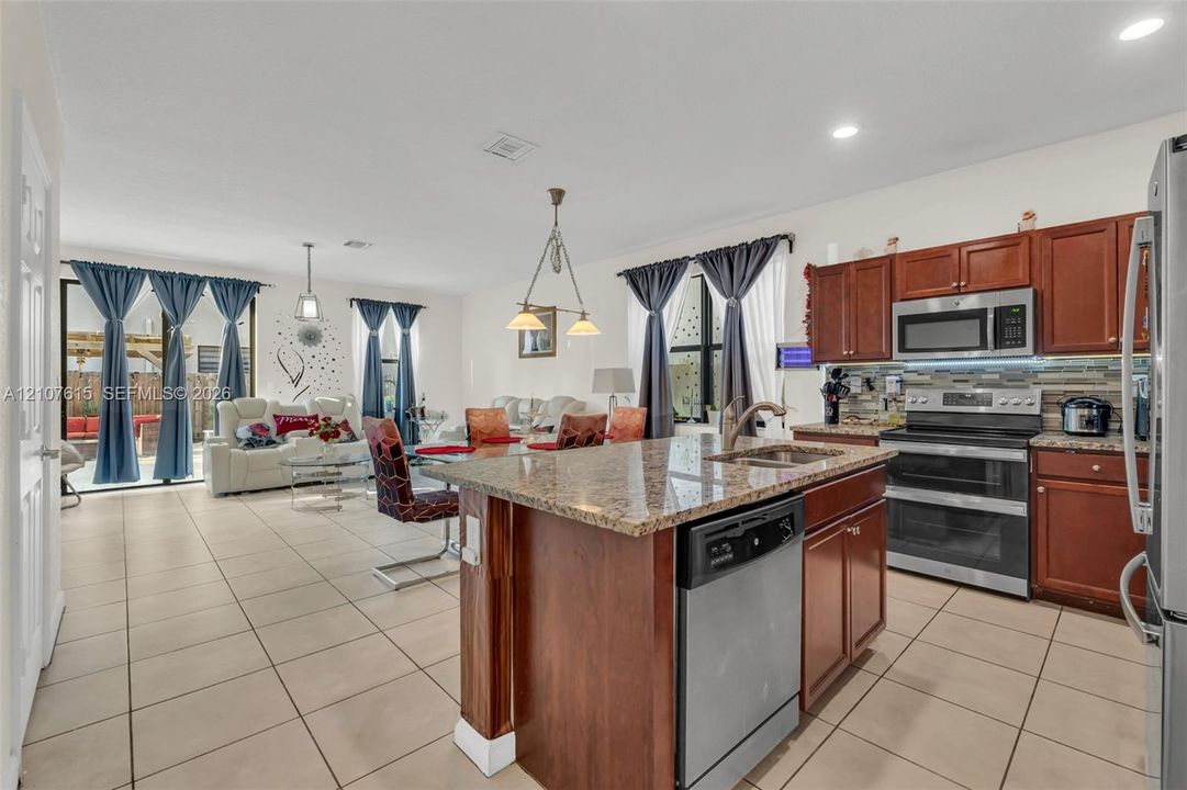Open-concept kitchen featuring an island with stone-finish countertops, warm-tone wooden cabinetry, stainless steel appliances, and neutral tile flooring throughout