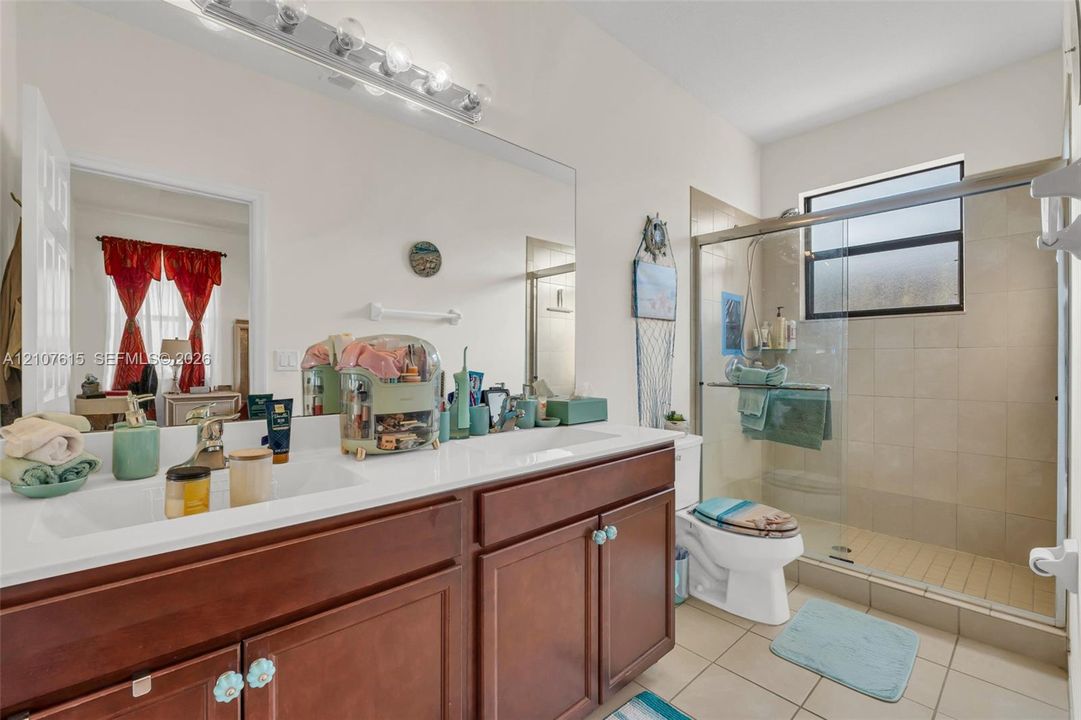 Bathroom featuring a dual-sink vanity with a solid-surface countertop, full-width wall mirror, wood-finish cabinetry, and a walk-in shower with tile surround and glass enclosure