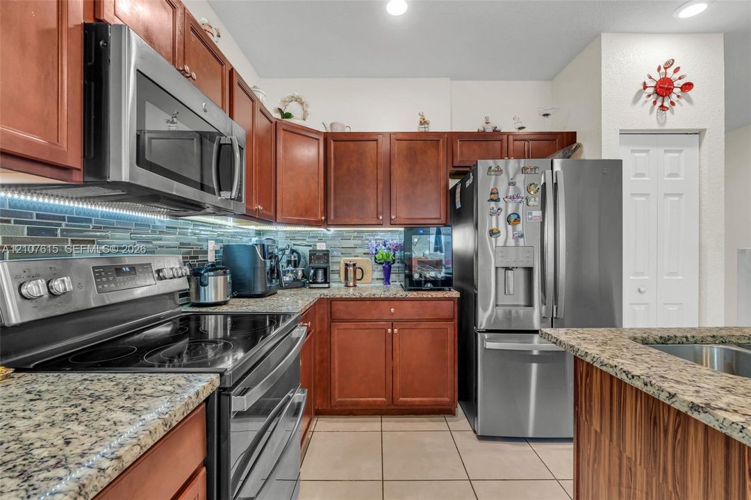 Kitchen featuring rich-toned wood cabinetry, stone-finish countertops, stainless steel appliances, multi-tonal glass tile backsplash, and ceramic floor tiling