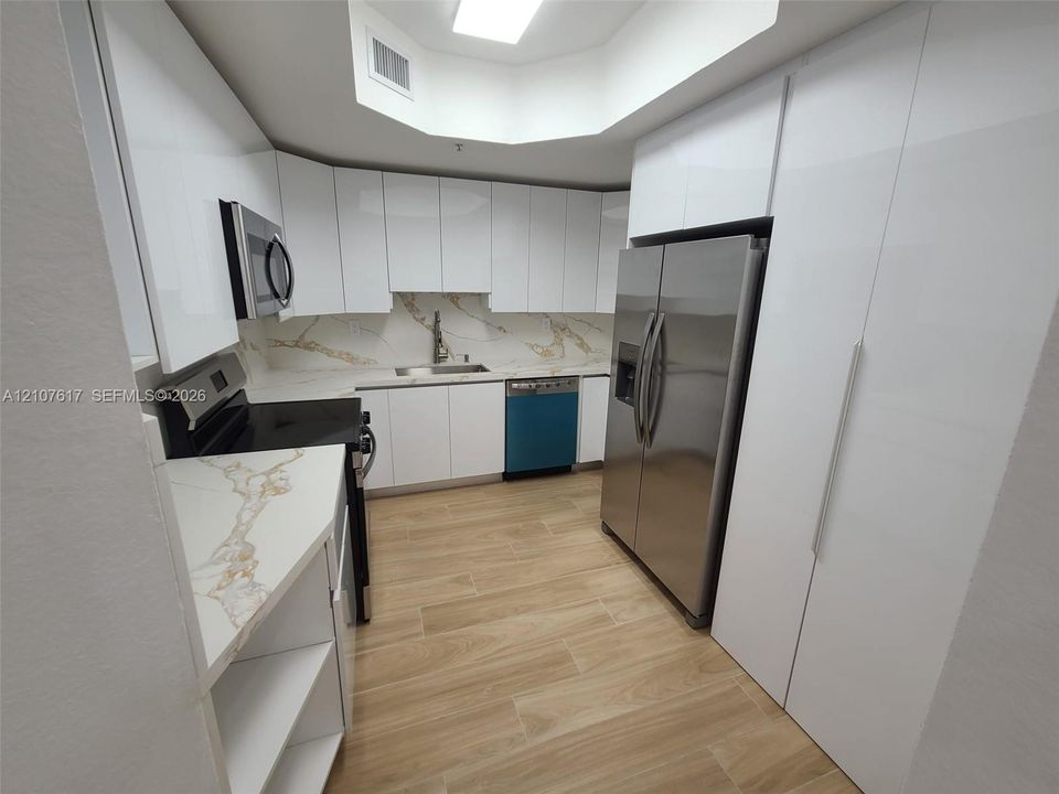 Modern kitchen featuring high-gloss white cabinetry, stone-finish countertops and backsplash, integrated stainless steel appliances, and wood-finish flooring under a recessed ceiling light fixture
