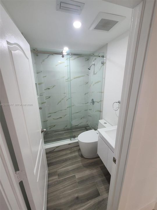 Walk-in shower with floor-to-ceiling stone-pattern tile, frameless glass sliding door, wood-finish flooring, and white floating vanity