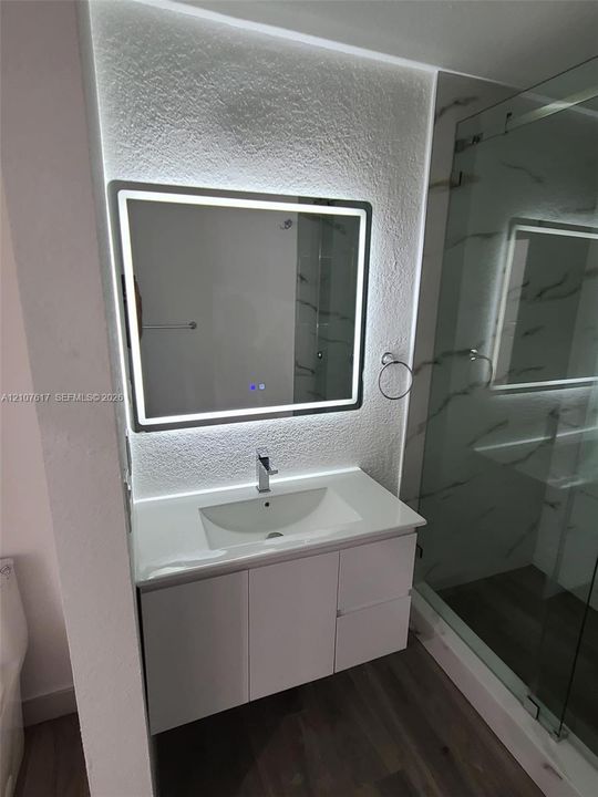 Floating vanity with white countertop and integrated sink, illuminated rectangular LED mirror, textured wall finish, and glass-enclosed shower with wall tile