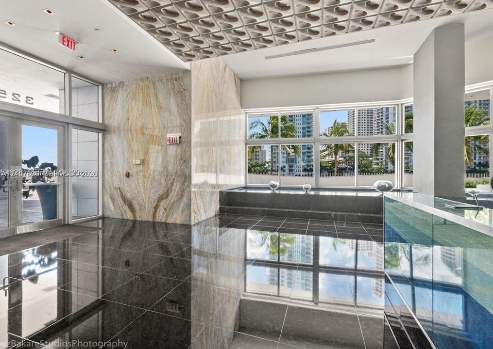 Lobby interior featuring large-format dark stone flooring, book-matched stone wall cladding, a geometric-patterned drop ceiling, floor-to-ceiling glass paneling, and a custom built-in reception desk