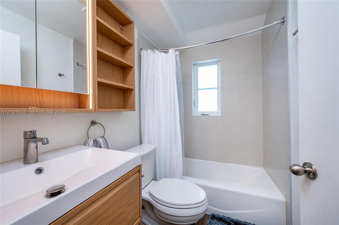 Wood-finish vanity with integrated rectangular sink and side shelving, wall-mounted lighted medicine cabinet, and combination tub and shower with light-toned wall tile