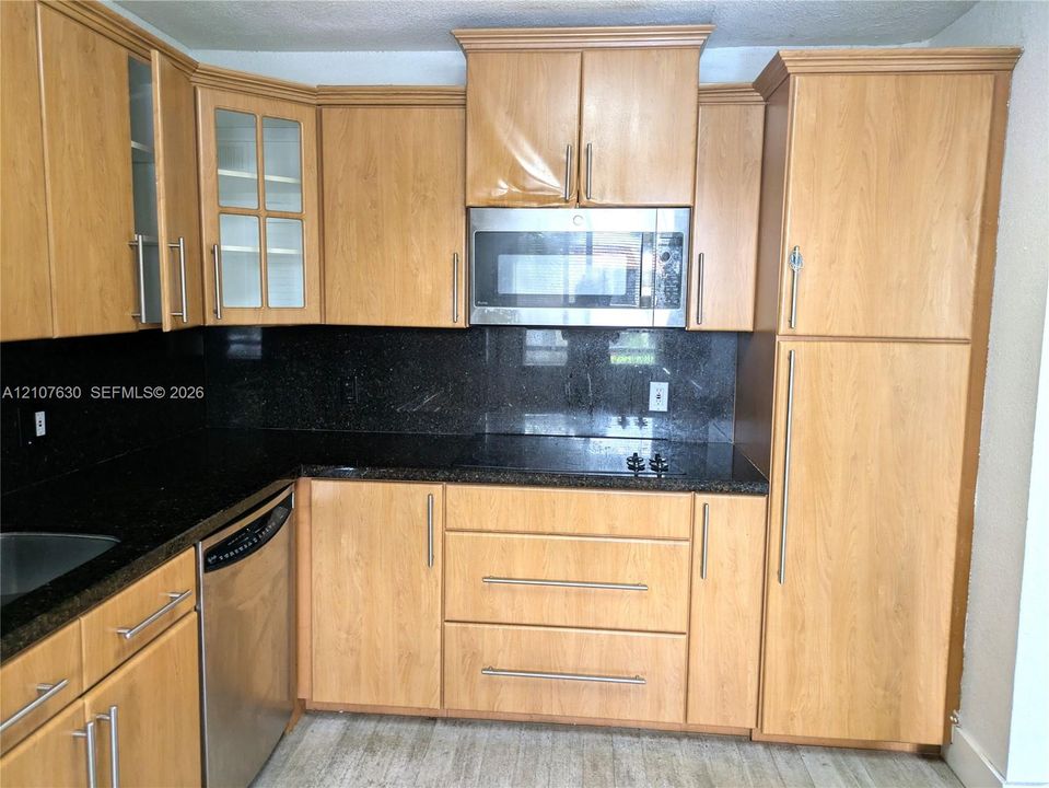 Kitchen featuring light-finish wood cabinetry, dark stone countertops, stainless steel dishwasher, built-in microwave, and glass-front upper cabinet doors