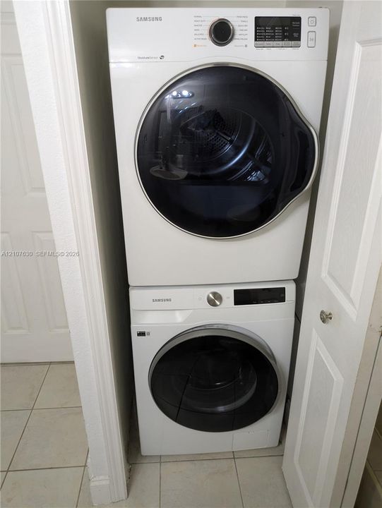 Stacked white front-load washer and dryer units with digital controls and glass-paneled doors, situated within a recessed laundry closet featuring tile flooring