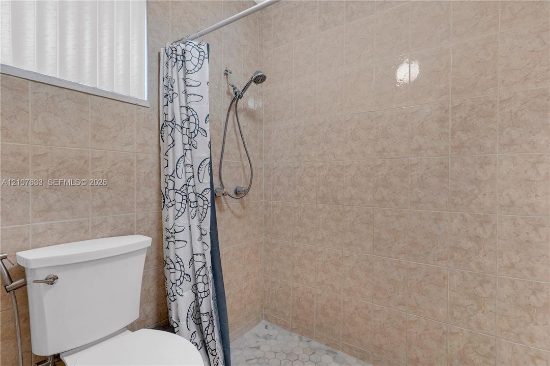 Shower stall featuring floor-to-ceiling tile, a handheld shower fixture, a white ceramic toilet, and a light-toned hexagonal tile floor