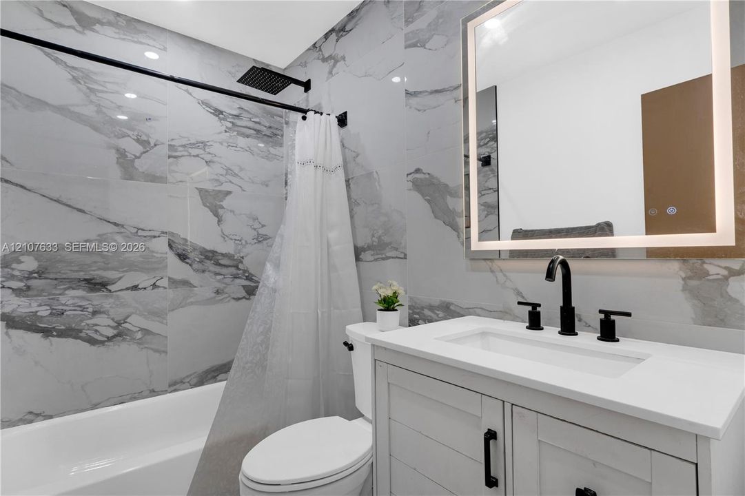 Bathroom featuring floor-to-ceiling large-format tile with veining, white vanity with stone-composite countertop, matte black widespread faucet, and LED-backlit mirror