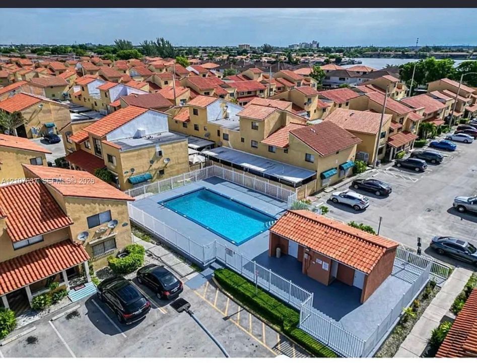 Aerial perspective of a community featuring a rectangular swimming pool, fenced pool deck, and a clubhouse structure with a clay tile roof