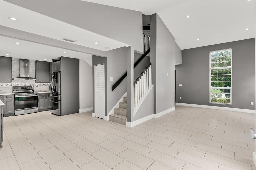Open-concept interior featuring vaulted ceilings, recessed lighting, tile flooring, a staircase with a white spindle railing, and a kitchen equipped with stainless steel appliances and custom cabinetry
