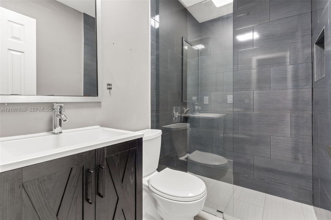 Bathroom featuring a vanity with dark wood-finish cabinetry, a glass-enclosed shower with charcoal-toned tile surround, built-in shower niche, and rectangular wall mirror