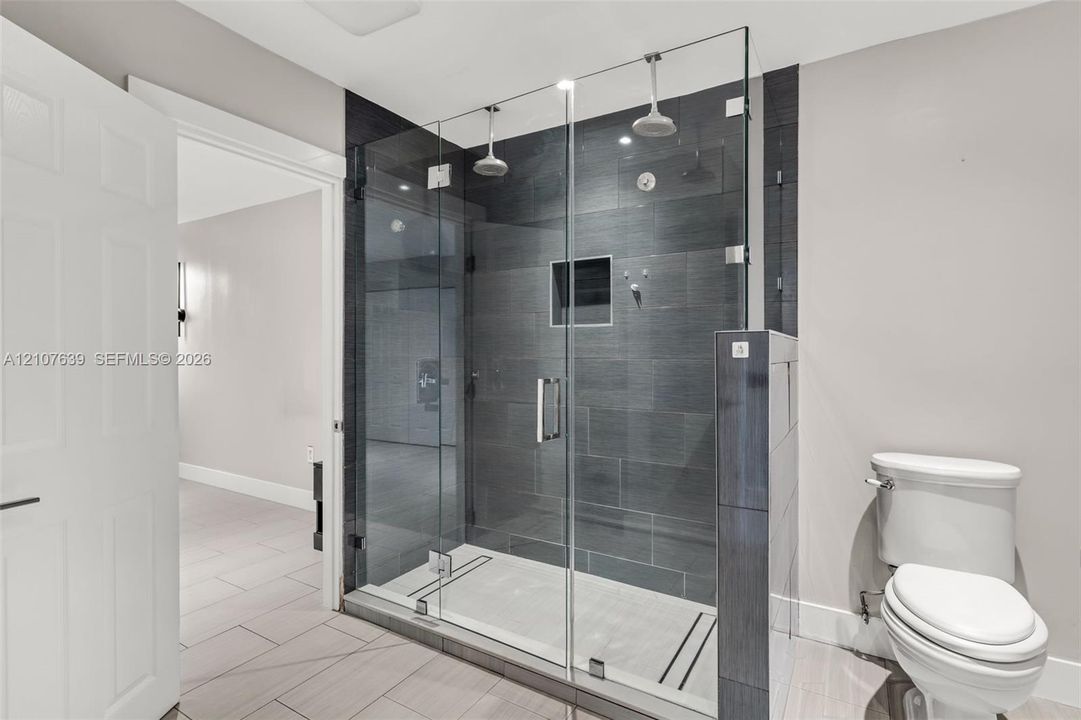 Walk-in shower featuring a glass enclosure, dual overhead showerheads, recessed wall niche, and dark large-format tile surround