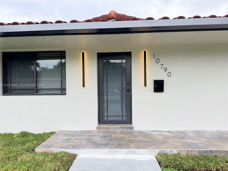 Exterior entryway featuring a terracotta tile roof, stone-finish porch, multi-pane glass-inset door, and vertical linear wall-mounted lighting