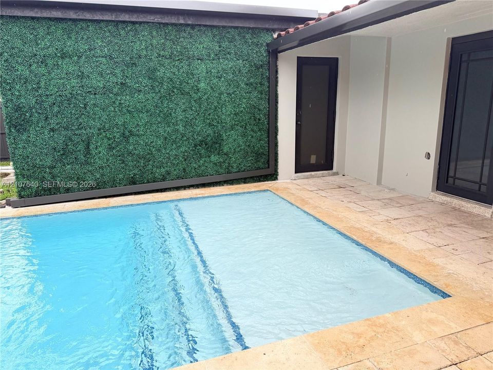 Swimming pool with a stone paver deck, artificial greenery wall feature, and black-framed glass entry doors