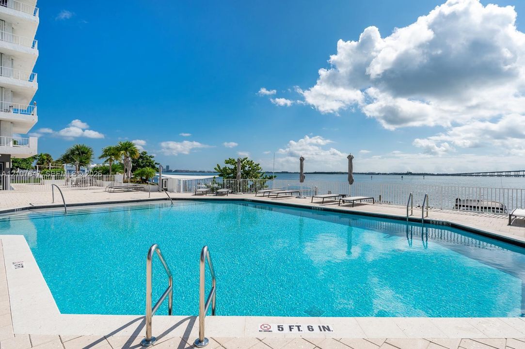 Swimming pool featuring metal handrails, stone paver decking, and direct water frontage with a protective perimeter fence