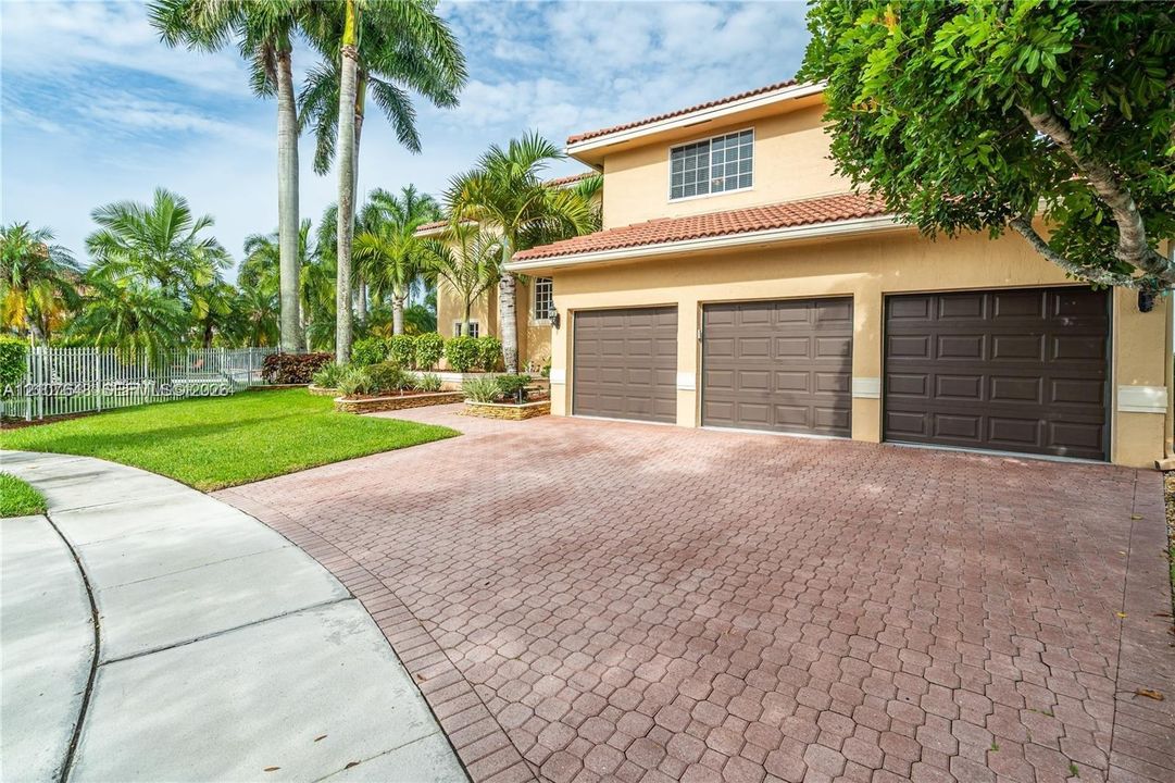 Three-car garage with paneled doors, expansive brick paver driveway, stucco exterior, clay tile roof, and manicured landscaping