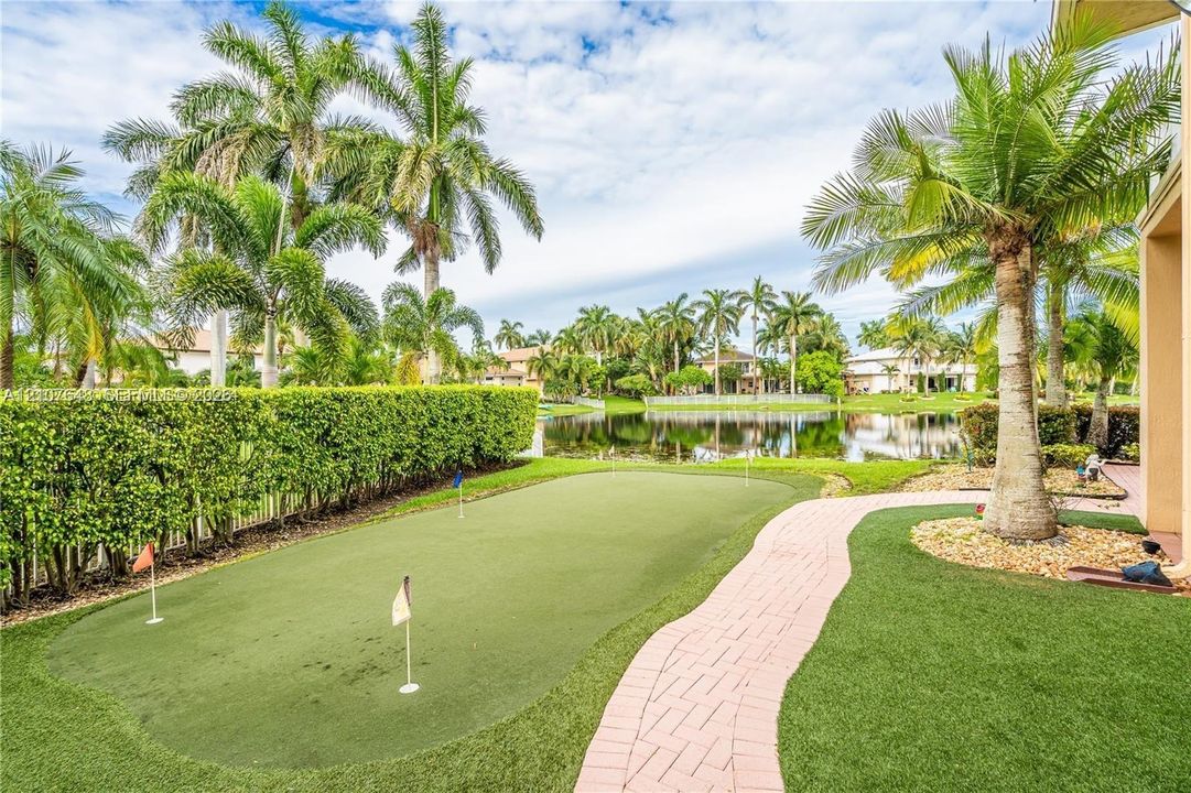 Backyard featuring a synthetic putting green, brick paver walkway, mature palm trees, privacy hedging, and water frontage