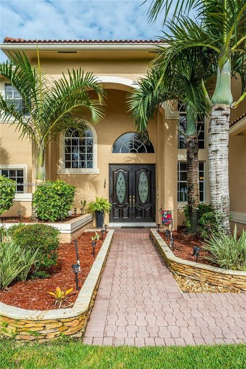 Stucco exterior featuring double entry doors with decorative glass insets, arched transom window, stacked stone landscape planters, and paved walkway