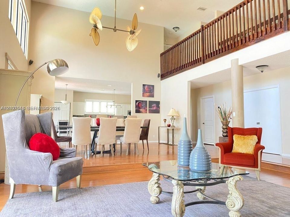 Double-height ceilings, elevated dining area, mezzanine balcony with wood railing, polished wood-finish flooring, and multi-blade ceiling fan