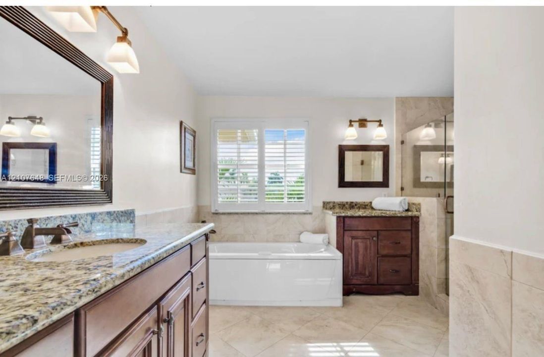 Bathroom featuring dual wood-finish vanities with stone countertops, an inset soaking tub, tile flooring, and shuttered windows