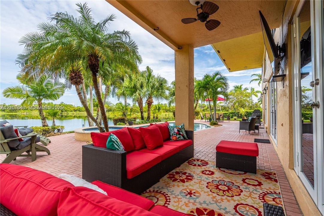 Covered patio with brick-style pavers, ceiling fan, built-in wall-mounted television, and direct access to an in-ground swimming pool overlooking a lake