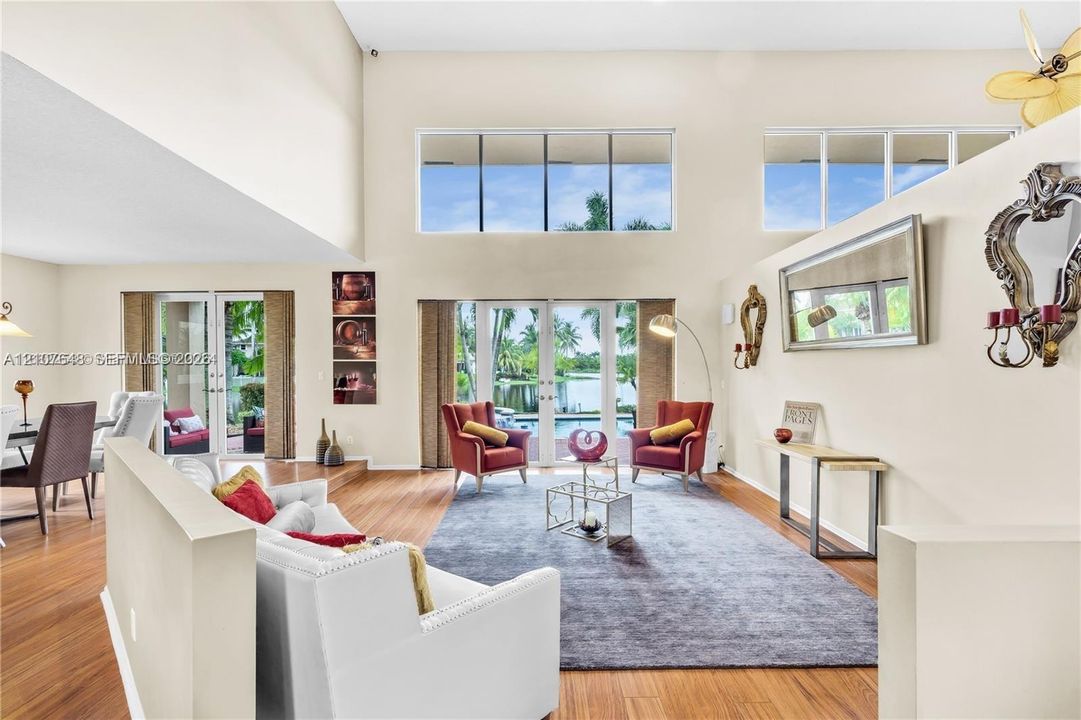 Expansive living area featuring vaulted ceilings, double-height windows, wood-finish flooring, and dual glass-paned doors with exterior water scenery