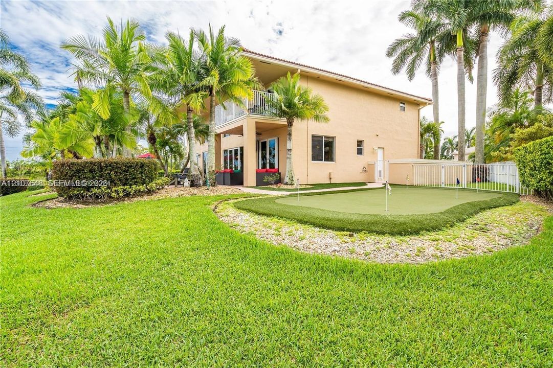 Rear exterior featuring a two-story structure, synthetic putting green, manicured lawn, established palm trees, and an upper-level balcony
