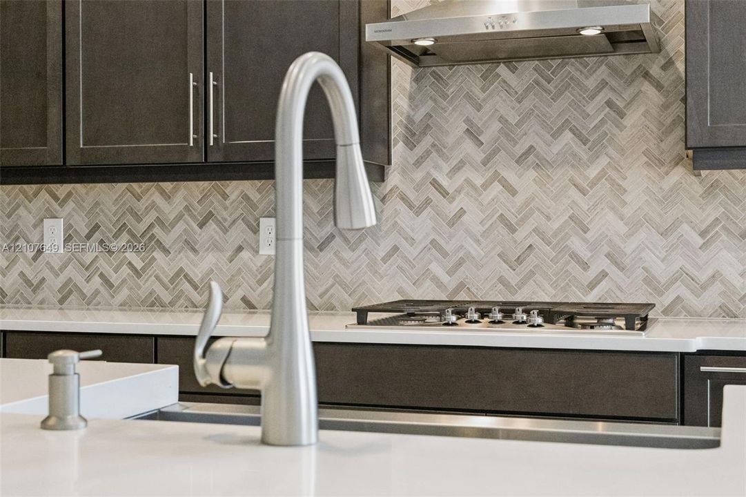 Kitchen featuring dark cabinetry, a herringbone-pattern backsplash, solid-surface countertops, a stainless steel gas cooktop, and a high-arc pull-down faucet