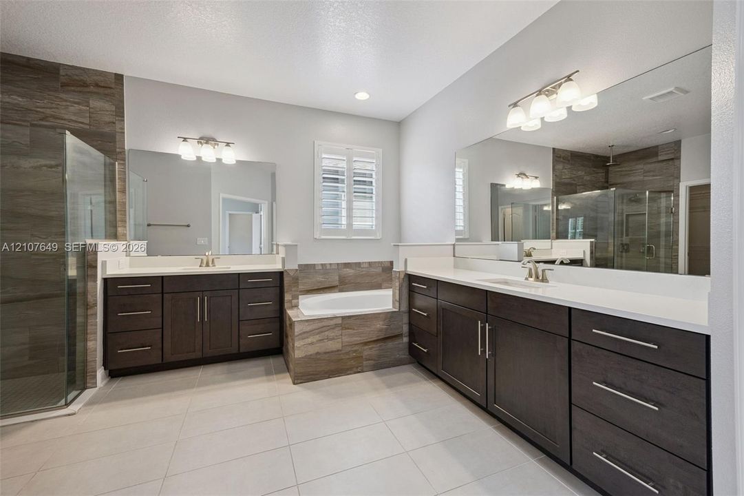 Dual vanities featuring dark cabinetry, solid-surface countertops, and expansive wall-to-wall mirrors, paired with a glass-enclosed shower and a deep soaking tub accented with earth-tone tile surrounds