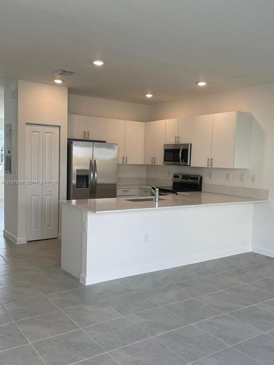 Kitchen featuring white flat-panel cabinetry, a stainless steel refrigerator and microwave, a peninsula with stone-surface countertops, and gray tile flooring