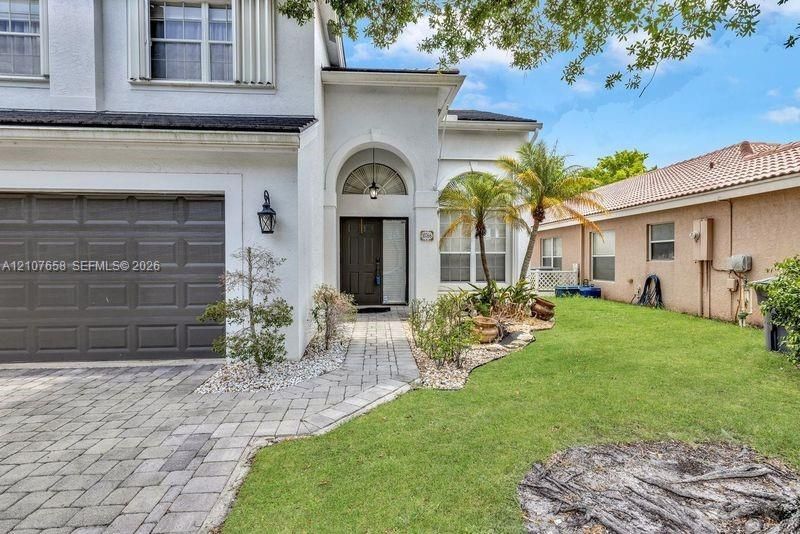 Stucco exterior featuring an arched entryway, paver driveway, dark-toned garage door, and a manicured lawn with mature palm landscaping