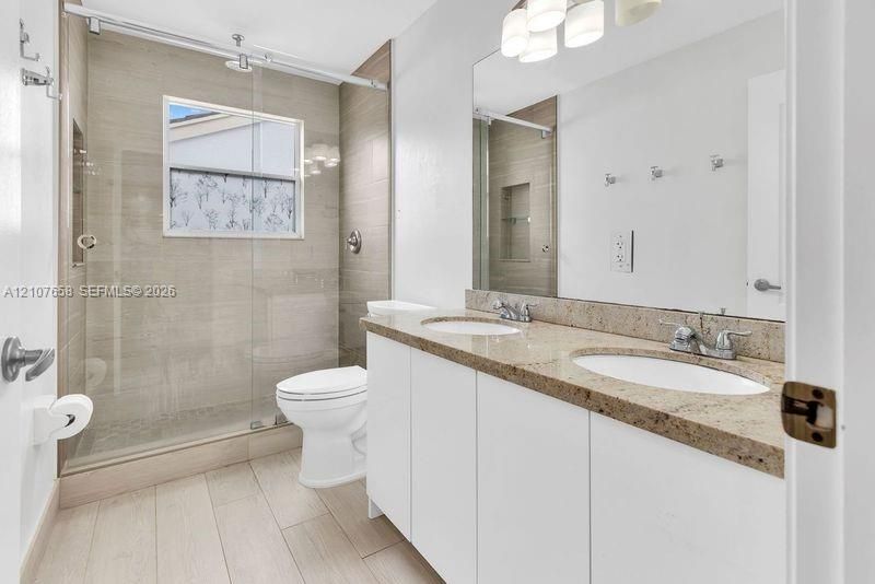 Dual-sink vanity with stone countertop, walk-in shower featuring glass sliding doors and tiled surround, and wood-finish flooring