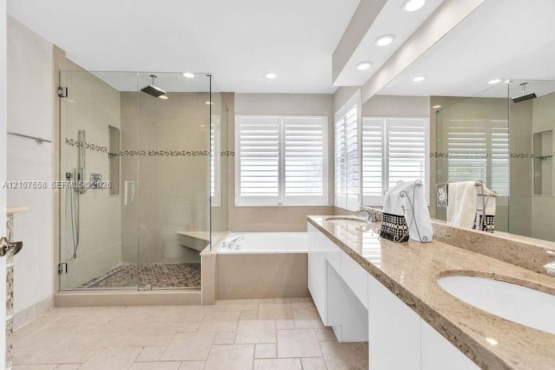 Bathroom featuring a dual-sink vanity with stone countertops, a glass-enclosed walk-in shower with a tiled bench and pebble-style flooring, a deep soaking tub, and custom plantation shutters